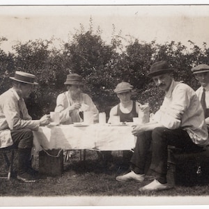 Pode incluir: Fotografia vintage em preto e branco de cinco pessoas sentadas à volta de uma mesa ao ar livre. Estão vestidas com camisas claras, chapéus e calças, desfrutando de uma refeição ou chá. A mesa está coberta com uma toalha branca.
