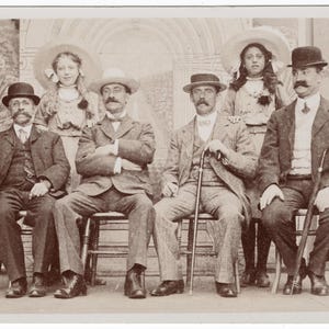 May include: Sepia-toned vintage photograph featuring seven people, four men seated in chairs and three women standing behind them. The men wear suits and hats, some with canes. The women wear hats and dresses. The background is a painted backdrop.