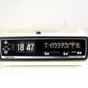 Puede incluir: Radio reloj Siemens Alpha vintage color crema que muestra las 18:47. Cuenta con un panel de visualización negro con indicadores de frecuencia de radio y diales de sintonización. El reloj tiene una parte superior texturizada y un cable de alimentación.