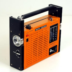 Peut inclure: Radio vintage orange Sony Sports 11 avec des détails noirs. Comprend une poignée, un bouton de réglage et une grille de haut-parleur. Le radio porte l'inscription "SONY SOLID STATE" et "SPORTS 11".
