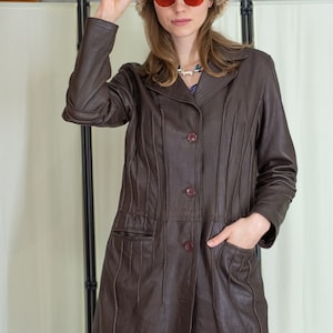 Puede incluir: Una chaqueta de cuero marrón con una silueta larga y ajustada. La chaqueta tiene un cuello, un cierre de botones y dos bolsillos. La modelo lleva un sombrero de piel marrón y gafas de sol rojas.