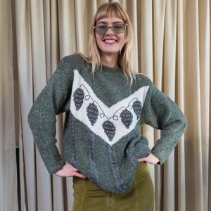 Può includere: Una donna indossa un maglione verde con un design geometrico bianco e grigio e una gonna di velluto a coste verde. Il maglione ha un motivo di pigne e foglie.