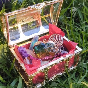 May include: A gold treasure chest with a red lining is open and filled with crystals and a heart-shaped pendant. The chest is sitting on a bed of green grass.