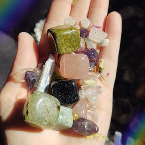 May include: A hand holding a variety of tumbled stones and crystals, including green aventurine, rose quartz, black obsidian, and amethyst.