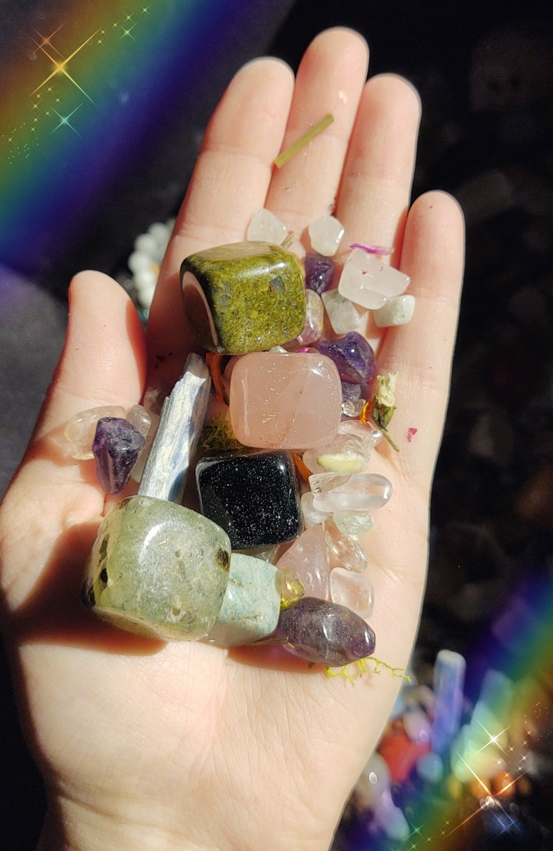 May include: A hand holding a variety of tumbled stones and crystals, including green aventurine, rose quartz, black obsidian, and amethyst.