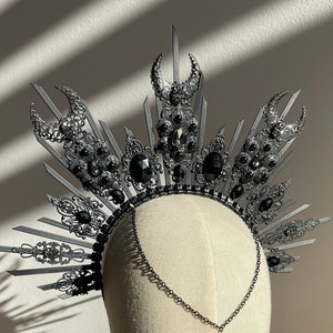 May include: A silver and black crown with a black teardrop pendant. The crown is made of metal and has a sunburst design with black crystals.