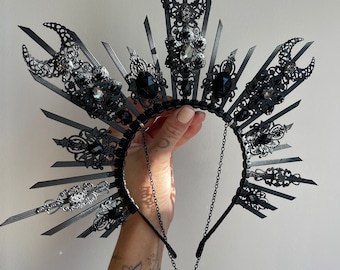 May include: A black headpiece featuring a sunburst design with pointed spikes, crescent moon motifs, and ornate filigree accents. The structure is embellished with faceted black and clear crystals and includes a delicate black chain with a teardrop pendant.