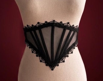 May include: A black waist corset featuring sheer mesh panels and vertical boning channels. The garment is finished with intricate black lace trim along the top and bottom edges, creating a pointed V-shape at the center front and back.