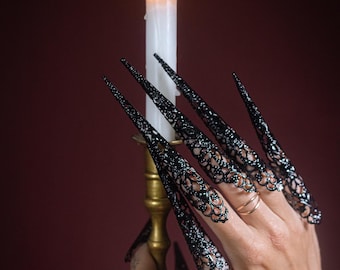 Op de afbeelding: Een hand met lange, zwarte, met kant bedekte vingernagels houdt een gouden kandelaar vast met een brandende witte kaars.