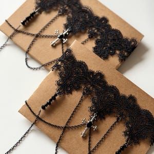 May include: Two black lace chokers featuring intricate floral patterns, delicate black metal chains, and small black beads. Each choker is adorned with a silver-toned cross pendant containing a central black stone, creating a layered design.