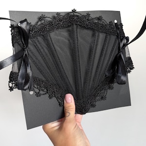 May include: A black corset-style accessory featuring sheer mesh panels and intricate lace trim along the top and bottom edges. The piece includes vertical boning channels and is secured on both sides with black satin ribbon ties for closure.