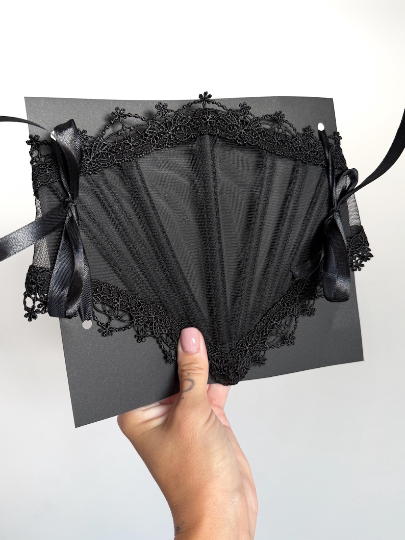 May include: A black corset-style accessory featuring sheer mesh panels and intricate lace trim along the top and bottom edges. The piece includes vertical boning channels and is secured on both sides with black satin ribbon ties for closure.