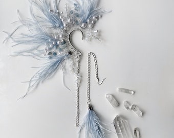May include: A light blue feather ear cuff with pearls, crystals, and a chain with a feather tassel. Several clear quartz crystals are scattered nearby. The ear cuff is designed to be worn on the ear.
