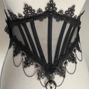May include: A black corset belt featuring sheer mesh panels and vertical boning. The edges are trimmed with intricate black lace. The bottom is adorned with delicate black chains and a central crescent moon charm with a small red gemstone.