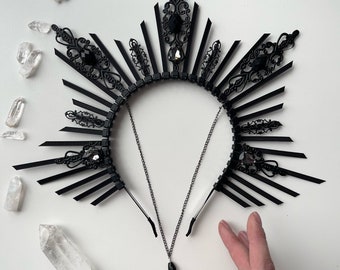 May include: A black metal crown with a sunburst design and black crystal accents. The crown is on a white background.