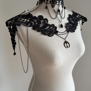 May include: A black lace choker and shoulder piece featuring intricate floral embroidery, delicate silver-toned chains, and dangling black teardrop beads. A central crescent moon pendant with a small red stone hangs from the layered chain design.