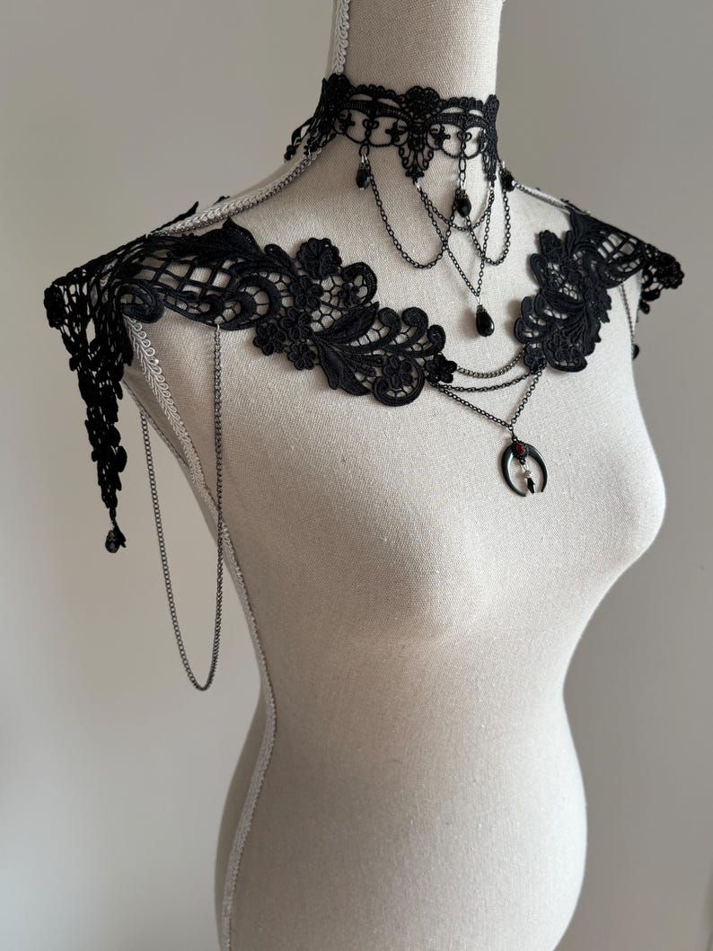 May include: A black lace choker and shoulder piece featuring intricate floral embroidery, delicate silver-toned chains, and dangling black teardrop beads. A central crescent moon pendant with a small red stone hangs from the layered chain design.