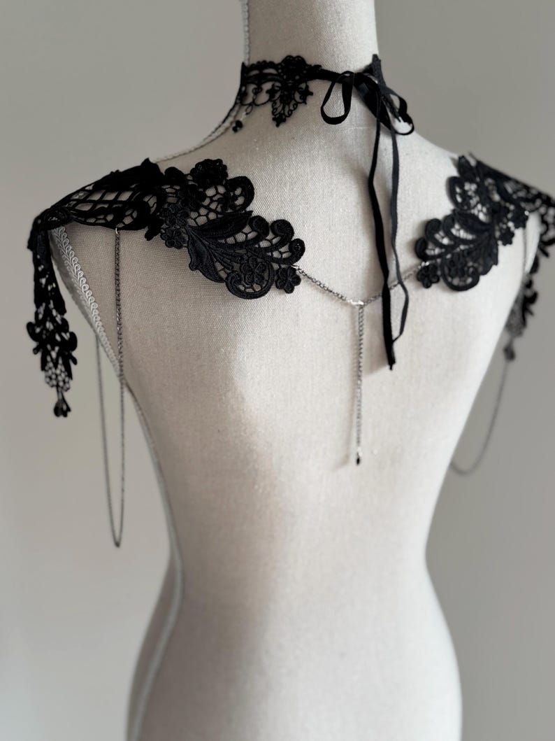 May include: A black lace shoulder accessory featuring intricate floral patterns, a matching choker collar with ribbon ties, and delicate silver-toned metal chains that drape across the back and shoulders, creating a layered and ornate appearance.