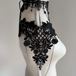 May include: A black lace choker and shoulder piece featuring intricate floral embroidery and scalloped edges. The accessory includes delicate silver-toned chains that connect the choker to the shoulder panels, accented with small black beads.
