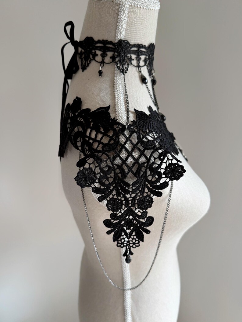 May include: A black lace choker and shoulder piece featuring intricate floral embroidery and scalloped edges. The accessory includes delicate silver-toned chains that connect the choker to the shoulder panels, accented with small black beads.