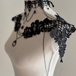 May include: A black lace choker and shoulder piece set featuring intricate floral patterns, delicate silver-toned chains, and small black beads. The choker includes a central pendant with a red accent, while the shoulder piece extends outward.