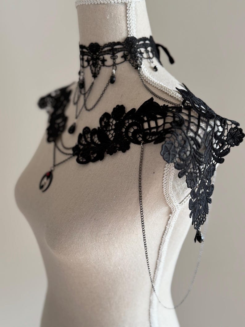 May include: A black lace choker and shoulder piece set featuring intricate floral patterns, delicate silver-toned chains, and small black beads. The choker includes a central pendant with a red accent, while the shoulder piece extends outward.