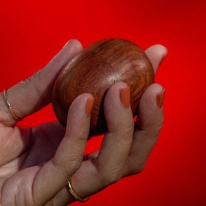 May include: A smooth, oval-shaped wooden object with a rich, dark brown color. The object is held in a hand against a vibrant red background. The wood grain is visible, adding texture to the piece.