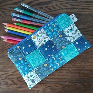 May include: A rectangular fabric pencil case featuring a patchwork design of blue and teal squares with various patterns including moons, stars, buildings, and nature motifs. Several coloured pencils and pens are tucked inside the open zip.