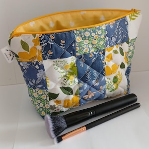 May include: A quilted makeup bag with a patchwork design featuring yellow floral patterns and navy blue fabric. The bag has a yellow interior and a zip closure. Two black makeup brushes with white and pink bristles are in front of the bag.