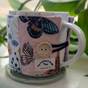 May include: A white ceramic mug with a quilted mug cosy featuring butterfly and floral patterns in pink, blue, and orange. The cosy is secured with a black elastic band and a wooden button that says "Handmade with love". The Patchwork Beagle label is visible.
