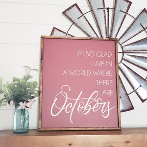 K&ouml;nnte beinhalten: Ein gerahmtes Schild mit einem malvenfarbenen Hintergrund und einem dunkelbraunen Holzrahmen. Der wei&szlig;e Text lautet: &bdquo;ICH BIN SO FROH, DASS ICH IN EINER WELT LEBE, IN DER ES Oktober gibt.&ldquo; Eine dekorative Windm&uuml;hle und ein blaues Einmachglas mit Blumen befinden sich im Hintergrund.