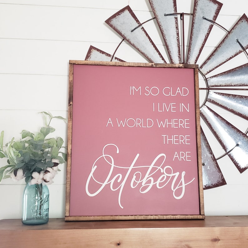 K&ouml;nnte beinhalten: Ein gerahmtes Schild mit einem malvenfarbenen Hintergrund und einem dunkelbraunen Holzrahmen. Der wei&szlig;e Text lautet: &bdquo;ICH BIN SO FROH, DASS ICH IN EINER WELT LEBE, IN DER ES Oktober gibt.&ldquo; Eine dekorative Windm&uuml;hle und ein blaues Einmachglas mit Blumen befinden sich im Hintergrund.