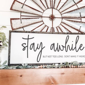 May include: A white wooden sign with black lettering that reads "stay awhile but not too long. don't make it weird". The sign is hanging on a wooden mantel with a metal windmill behind it.