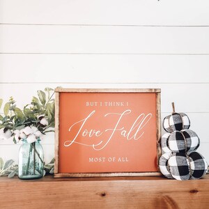 May include: A wooden framed sign with the text "BUT I THINK I Love Fall MOST OF ALL" in white script on an orange background. Beside the sign are fabric pumpkins in black and white buffalo check and a blue mason jar with cotton stems.