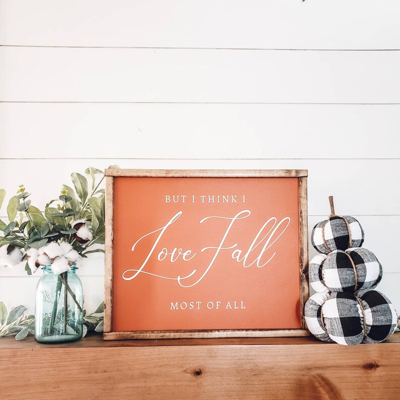 May include: A wooden framed sign with the text "BUT I THINK I Love Fall MOST OF ALL" in white script on an orange background. Beside the sign are fabric pumpkins in black and white buffalo check and a blue mason jar with cotton stems.
