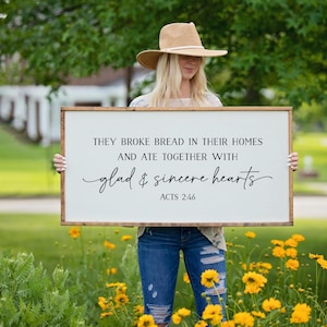 May include: White wooden sign with the text "They broke bread in their homes and ate together with glad & sincere hearts Acts 2:46". A woman is holding the sign in front of a green grassy background with yellow flowers.