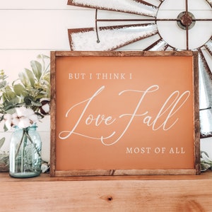 May include: A framed sign with the text "But I think I Love Fall Most of All" in white script on an orange background. The sign has a dark brown wooden frame and is decorated with a mason jar and greenery.