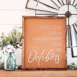 K&ouml;nnte beinhalten: Ein Holzschild mit dem Text "I'M SO GLAD I LIVE IN A WORLD WHERE THERE ARE Octobers" in wei&szlig;er Schrift auf orangefarbenem Hintergrund. Das Schild steht auf einer Holzoberfl&auml;che mit einer rustikalen Windm&uuml;hle und einem Einmachglas mit Blumen.