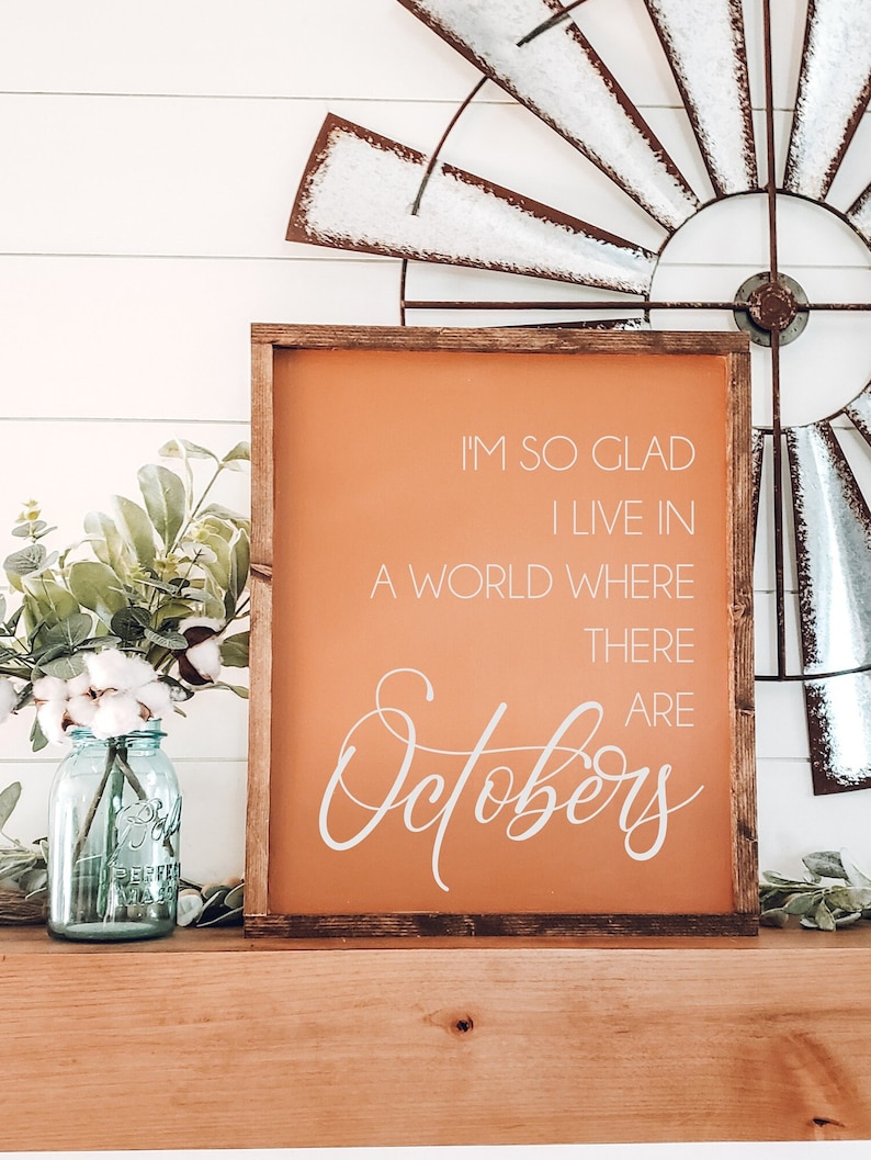 K&ouml;nnte beinhalten: Ein Holzschild mit dem Text "I'M SO GLAD I LIVE IN A WORLD WHERE THERE ARE Octobers" in wei&szlig;er Schrift auf orangefarbenem Hintergrund. Das Schild steht auf einer Holzoberfl&auml;che mit einer rustikalen Windm&uuml;hle und einem Einmachglas mit Blumen.