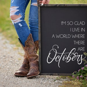 K&ouml;nnte beinhalten: Ein schwarzes Schild mit wei&szlig;em Text: &bdquo;I'M SO GLAD I LIVE IN A WORLD WHERE THERE ARE Octobers&ldquo;. Die Person tr&auml;gt blaue Jeans und braune Cowboy-Stiefel.
