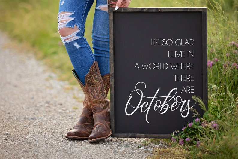 K&ouml;nnte beinhalten: Ein schwarzes Schild mit wei&szlig;em Text: &bdquo;I'M SO GLAD I LIVE IN A WORLD WHERE THERE ARE Octobers&ldquo;. Die Person tr&auml;gt blaue Jeans und braune Cowboy-Stiefel.