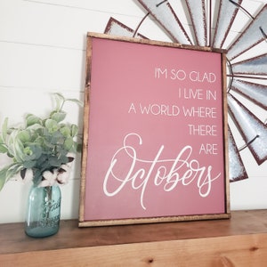 K&ouml;nnte beinhalten: Ein gerahmtes Schild mit einem staubrosa Hintergrund und dem Text "I'M SO GLAD I LIVE IN A WORLD WHERE THERE ARE Octobers" in wei&szlig;er Schrift. Der Rahmen ist dunkelbraun. Links steht ein blaues Einmachglas mit Gr&uuml;n und Baumwollstielen.
