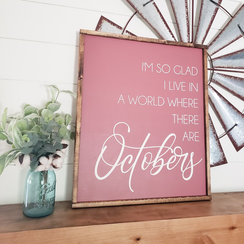 K&ouml;nnte beinhalten: Ein gerahmtes Schild mit einem staubrosa Hintergrund und dem Text "I'M SO GLAD I LIVE IN A WORLD WHERE THERE ARE Octobers" in wei&szlig;er Schrift. Der Rahmen ist dunkelbraun. Links steht ein blaues Einmachglas mit Gr&uuml;n und Baumwollstielen.