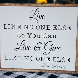 May include: A white wooden sign with a black inscription that reads "Live like no one else so you can live & give like no one else - Dave Ramsey".