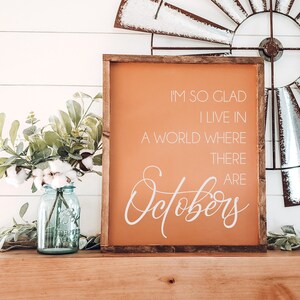 K&ouml;nnte beinhalten: Ein gerahmtes Schild mit orangefarbenem Hintergrund und wei&szlig;em Text: "I'M SO GLAD I LIVE IN A WORLD WHERE THERE ARE Octobers". Links steht ein blaues Glasgef&auml;&szlig; mit Baumwolle und Gr&uuml;nzeug.