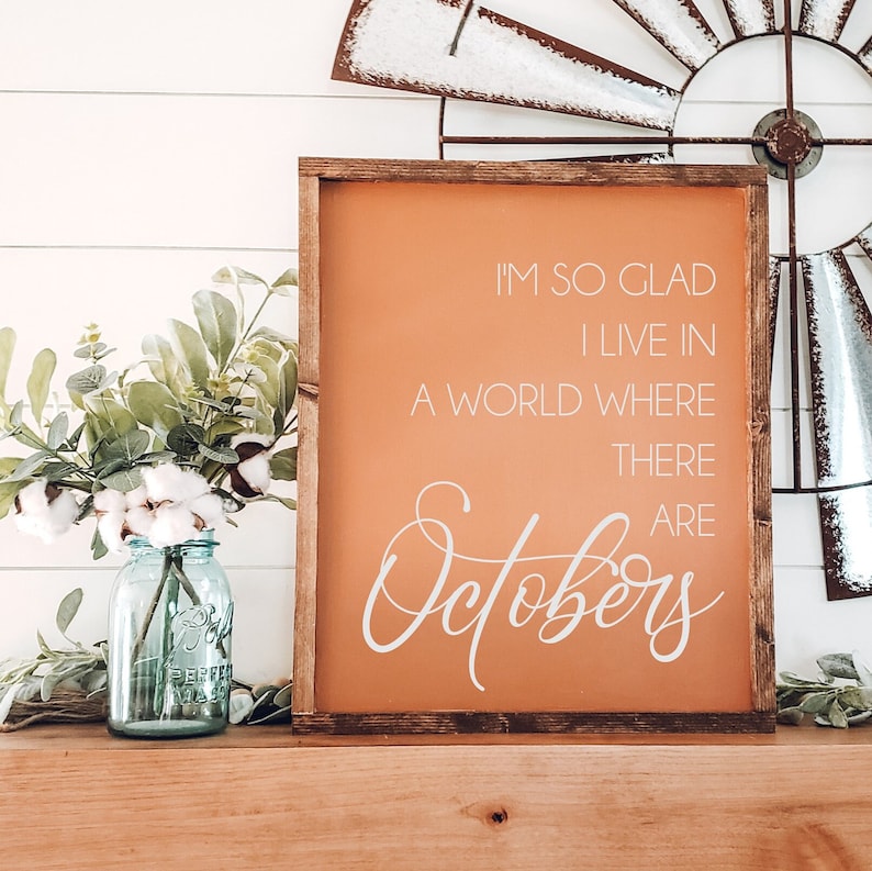 K&ouml;nnte beinhalten: Ein gerahmtes Schild mit orangefarbenem Hintergrund und wei&szlig;em Text: "I'M SO GLAD I LIVE IN A WORLD WHERE THERE ARE Octobers". Links steht ein blaues Glasgef&auml;&szlig; mit Baumwolle und Gr&uuml;nzeug.