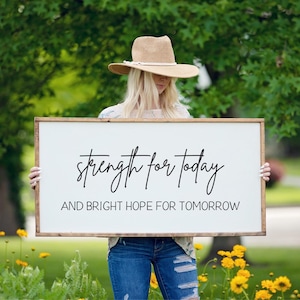May include: A rectangular wooden sign with a white background and a brown frame. The sign displays the text "strength for today" and "AND BRIGHT HOPE FOR TOMORROW" in black script. The sign is held outdoors in front of a green background.