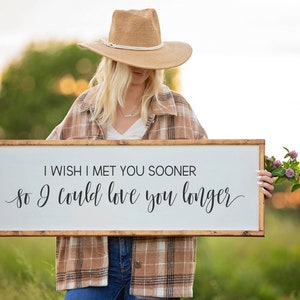 May include: A woman wearing a brown and white plaid shirt and a brown cowboy hat holds a wooden sign with the text "I wish I met you sooner so I could love you longer".