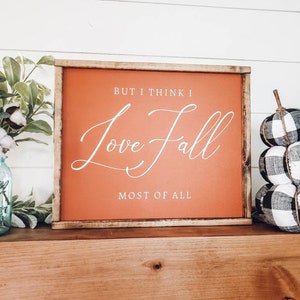 May include: A framed sign with the text "But I think I Love Fall Most of All" in white script on an orange background. Beside the sign are a blue mason jar with flowers and a stack of black and white plaid pumpkins.