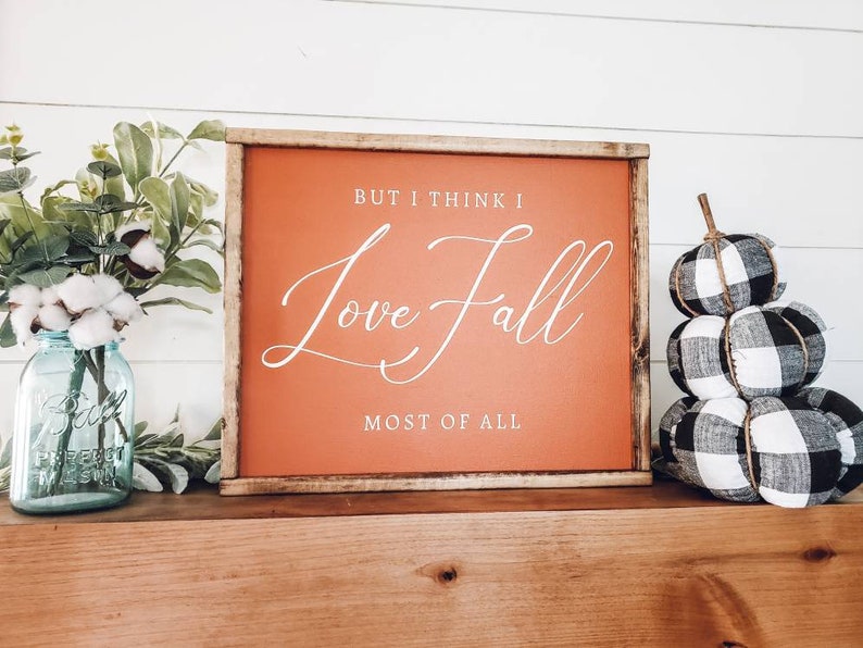 May include: A framed sign with the text "But I think I Love Fall Most of All" in white script on an orange background. Beside the sign are a blue mason jar with flowers and a stack of black and white plaid pumpkins.